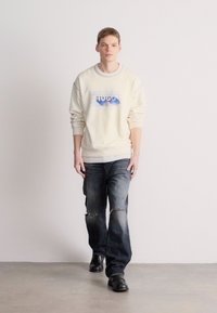 Krém színű pulóver kék "Hugo" logóval, sötét kék farmernadrággal, melyen kopott részek találhatók. A modell fekete cipőt visel; semleges háttér.