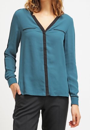 Türkise Bluse mit langen Ärmeln aus glattem Stoff, mit V-Ausschnitt, kontrastierendem schwarzen Saum und zwei kleinen Brusttaschen. Getragen mit schwarzen Hosen.