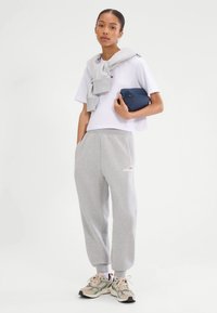 Kobieta w szarych dresach i białym krótki topie, trzymająca granatową saszetkę, z pasującą bluzą narzuconą na ramiona, stojąca.