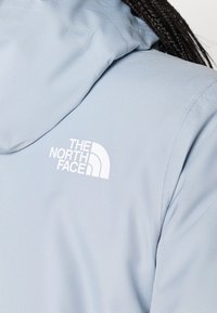 Ανοιχτό μπλε μπουφάν με υφασμάτινη υφή, με λευκό λογότυπο "The North Face" στο πάνω μέρος της πλάτης. Κουκούλα μερικώς ορατή.
