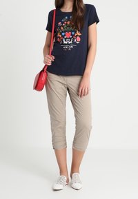 Marineblauw grafisch T-shirt met kleurrijke bloemenprint, beige cropped broek, witte instapschoenen en een rode crossbodytas.
