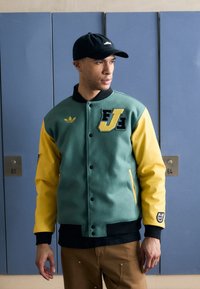 Green and yellow varsity jacket with embroidered letters, black collar, snap buttons, side pockets, and patch details on the sleeve.