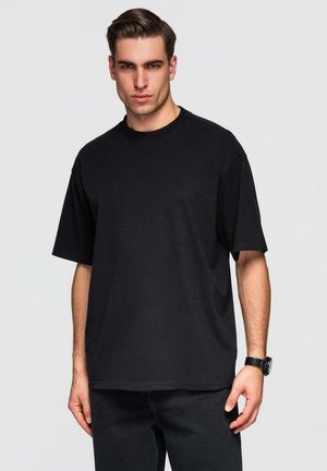 Ung mand med kort, tilbagestrøget hår iført en sort oversize T-shirt og sort ur, stående mod en ensfarvet, lys baggrund.