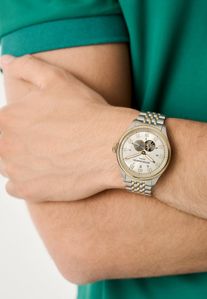 Reloj bicolor en plata y oro con una esfera redonda, esfera blanca y acentos negros. La pulsera presenta un diseño de eslabones. Mano descansando sobre el brazo.
