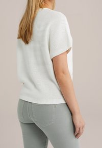 Žena s dlouhými blond vlasy, oblečená v bílém pleteném topu s krátkým rukávem a světle šedých úzkých kalhotách, zobrazená zezadu na béžovém pozadí.