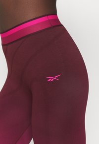 Zbliżenie na osobę o ciemnej karnacji, która nosi bordowe legginsy sportowe z różowym paskiem w talii oraz różowym logo Reebok na biodrze.