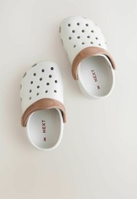 Sabots en caoutchouc blancs avec des sangles marron, dotés de trous de ventilation, d'un intérieur texturé et d'un nom de marque imprimé en rouge.
