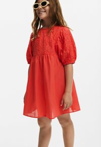 Röd broderad klänning med puffärmar och en figurnära livstycke, med en flödande kjol och textil med struktur. Modellen bär solglasögon.