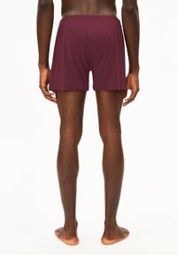 Shorts in cotone borgogna con vita elastica, lunghezza a metà coscia e superficie liscia, visti di spalle su piedi nudi.