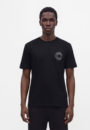Νέος άνδρας που φοράει μαύρο T-shirt Hugo Jeans με στρογγυλό λογότυπο στο στήθος, στέκεται μπροστά σε απλό λευκό φόντο.