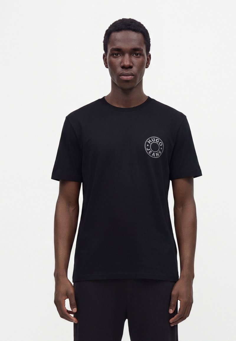 Νέος άνδρας που φοράει μαύρο T-shirt Hugo Jeans με στρογγυλό λογότυπο στο στήθος, στέκεται μπροστά σε απλό λευκό φόντο.