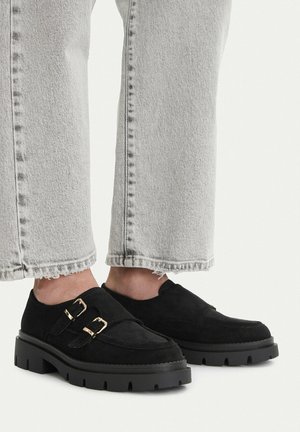 Mocassini neri in camoscio con doppia fibbia dorata indossati con jeans grigi chiari a orlo sfrangiato, mostrati dalla metà polpaccio in giù su sfondo bianco.