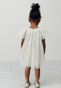 Robe courte en ivoire irisé avec des motifs d'étoiles, des manches bouffantes et un ourlet en forme de coquille. Associée à des chaussures argentées brillantes.