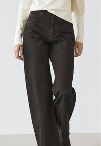 Zwarte jeans met wijde pijpen van denim stof, voorzien van een hoge taille, voorzakken en subtiele stiksel details. Gecombineerd met een lichtgekleurde top.