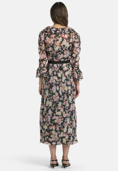 Femme aux longs cheveux vêtue d'une robe midi noire à motifs floraux avec des poignets à volants et une ceinture noire, debout de dos devant un fond uni.