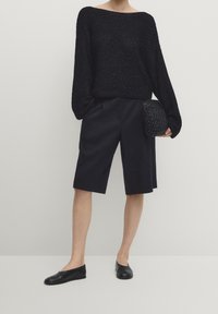 Maglione in maglia nera con texture, scollatura ampia, abbinato a pantaloni shorts neri sartoriali. Scarpe nere basse e una piccola borsa intrecciata.