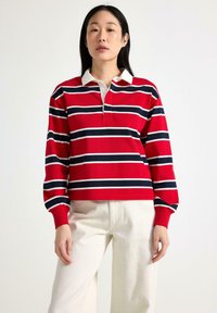 Rød langærmet polo-shirt med marineblå og hvide vandrette striber, med en hvid krave og knaplukning, parret med off-white bukser.