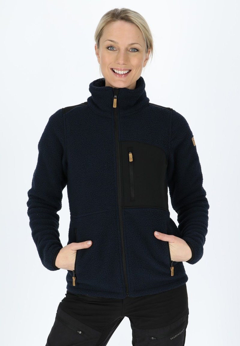 Navy fleece jacket with high collar, front zipper, black fabric pocket, and wooden accents. Features side pockets and textured material.