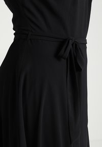 Svart klänning med en mjuk, flödande tyg och knytning i midjan. Designen inkluderar en subtil drapering och minimalistisk estetik.