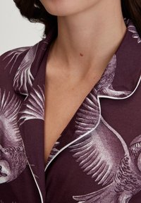 Pigiama color bordeaux con rifiniture bianche, caratterizzato da un motivo a uccelli. Il tessuto è morbido, con colletto classico e vestibilità rilassata.