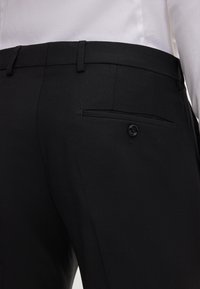 Schwarze, taillierte Hose mit glatter Textur, ausgestattet mit einer einzelnen Gesäßtasche mit Knopfverschluss und Gürtelschlaufen an der Taille.