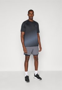 T-shirt de sport noir avec un motif de rayures bleues, assorti à un short gris. Le tissu semble respirant ; le logo Under Armour est présent sur le t-shirt.