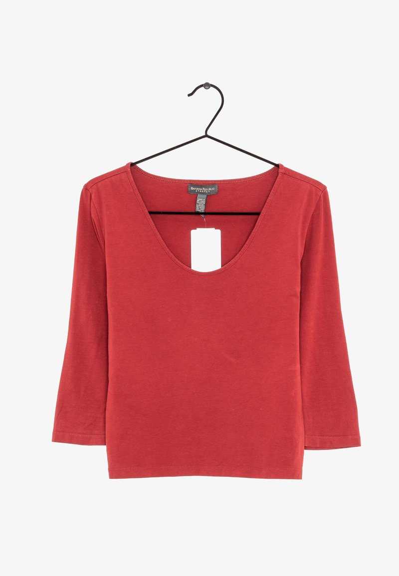 Červený top s dlouhým rukávem a hlubokým výstřihem ve tvaru V, vyrobený z měkkého materiálu. Obsahuje štítek visící z výstřihu. Jednoduchý design bez vzorů.