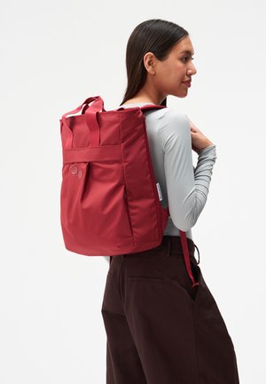 Frau mit großem rotem Rucksack, gekleidet in einem hellgrauen Langarmshirt und dunkelbraunen Hosen, seitlich auf einem einfarbigen weißen Hintergrund stehend.