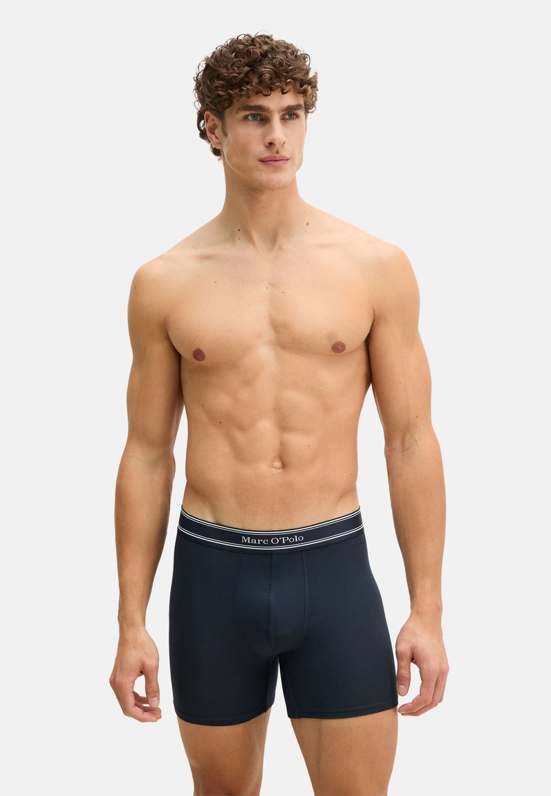 Homme torse nu aux cheveux bouclés portant un boxer Marc O’Polo bleu marine, debout devant un fond blanc uni.