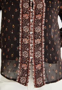 Blusa trasparente nera con motivi floreali e geometrici arancioni e gialli, dotata di chiusura con bottoni e dettagli in bottone bianchi.