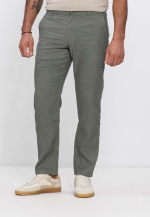 Mann trägt graue Straight-Fit-Hose und weiße Turnschuhe, steht vor einem schlichten hellen Hintergrund.