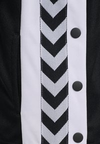 Schwarze Jacke mit einem weißen vertikalen Paneel mit Chevron-Muster. Verfügt über schwarze Druckknöpfe und eine glatte Stoffstruktur.
