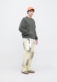 Felpa grigia con tasca frontale, pantaloni crema con macchie scure e camicia bianca annodata; cappellino arancione; scarpe grigie con suole marroni.