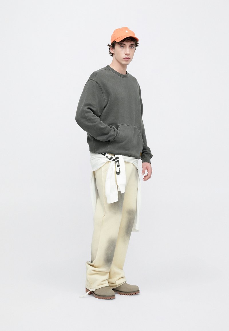 Grå sweatshirt med en framficka, krämfärgade byxor med mörka fläckar och en vit knuten skjorta; orange keps; grå skor med bruna sulor.