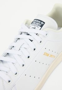 Hvid lædersneaker med tekstureret overflade, perforerede detaljer og guld "STAN SMITH" branding. Har flade hvide snørebånd og en beige sål.