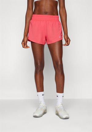 Femme portant un short de sport corail, des chaussettes blanches Nike et des chaussures de sport blanches, debout devant un fond clair uni.