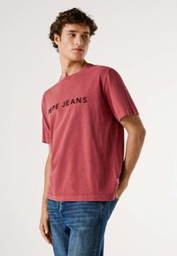 Camiseta roja de manga corta hecha de algodón con cuello redondo y logo negro "PEPE JEANS" en el pecho. Corte casual, dobladillo con abertura lateral.