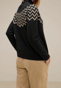 Person som bär en svart tröja med beige zigzag-mönster över axlarna, i kombination med beiga byxor, med händerna i fickorna, vänd bort.