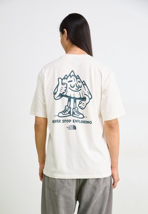 Person mit langen dunklen Haaren, die ein weißes T-Shirt mit einem Cartoon-Berg trägt, der Daumen hoch zeigt, und dem Text "Never Stop Exploring" auf dem Rücken.