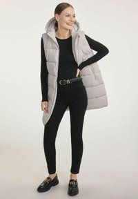Jasnoszara, ocieplana kamizelka bez rękawów z kapturem, zaprezentowana na czarnym, długim topie. Połączona z czarnymi spodniami i czarnymi butami z metalowymi akcentami.