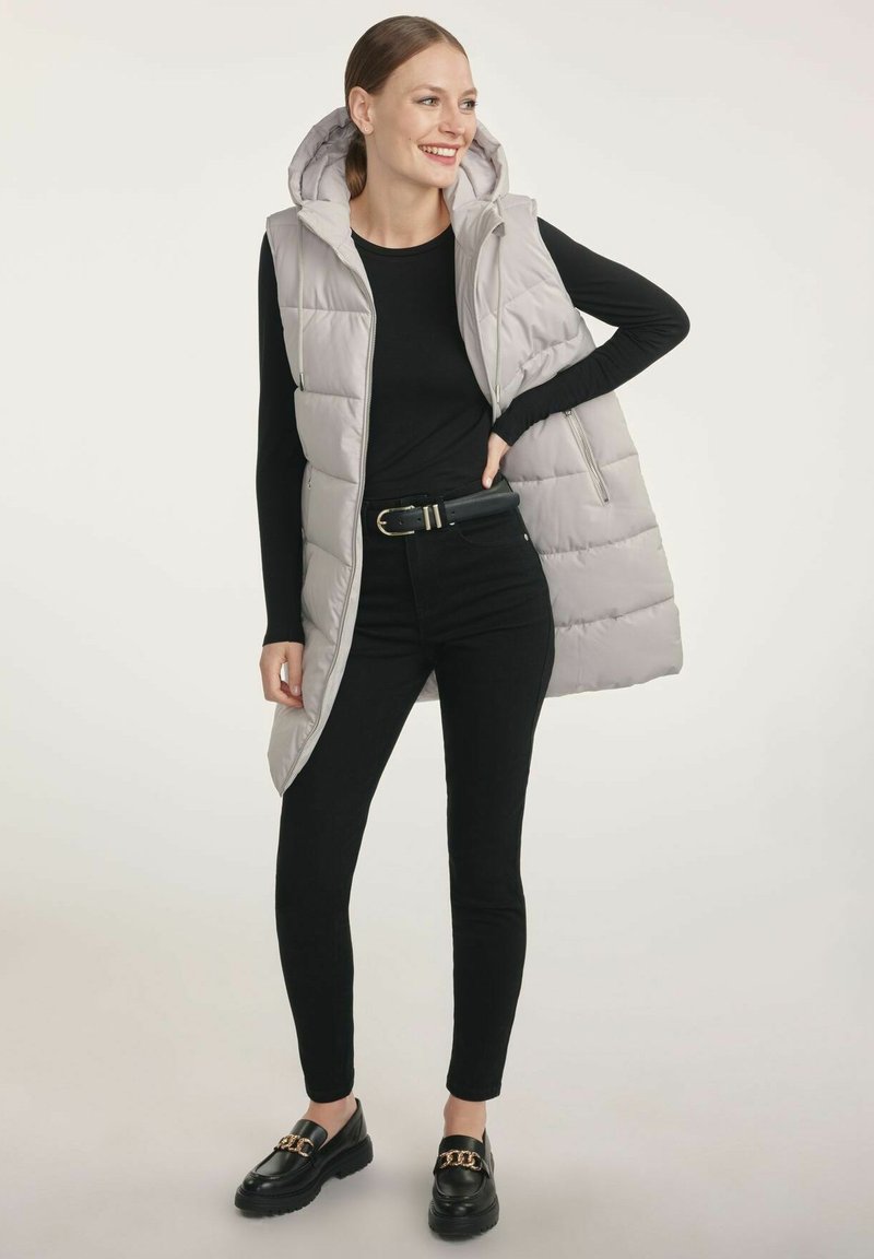 Jasnoszara, ocieplana kamizelka bez rękawów z kapturem, zaprezentowana na czarnym, długim topie. Połączona z czarnymi spodniami i czarnymi butami z metalowymi akcentami.