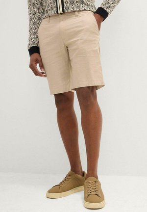 Shorts beige su misura realizzati in un tessuto leggero, con taglio dritto e tasche laterali, abbinati a sneakers beige.