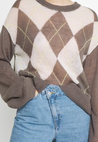 Brun og beige argyle-sweater med gul stikning, der har en afslappet pasform og ribbede manchetter, parret med højtaljede denimjeans.