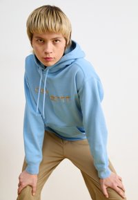 Hanorac albastru deschis cu sireturi și buzunar frontal, având logo-ul "Carhartt" în aur. Modelul poartă pantaloni bej, evidențiind o ținută casual.