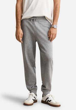 Homme portant un pantalon de jogging gris avec cordon, des baskets blanches avec des bandes noires, une chemise blanche et une montre-bracelet argentée, debout devant un fond blanc.