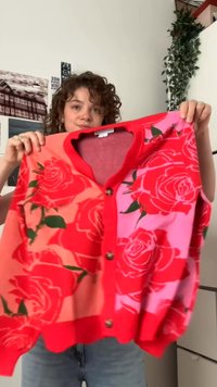 Woman with curly hair holding a red and pink floral button-up cardigan with large rose patterns in a room with wall photos and a hanging plant.