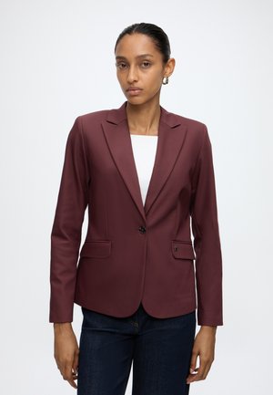 Frau trägt ein tailliertes bordeauxrotes Sakko über einem weißen Top und dunkelblauen Jeans, steht vor einem schlichten weißen Hintergrund.