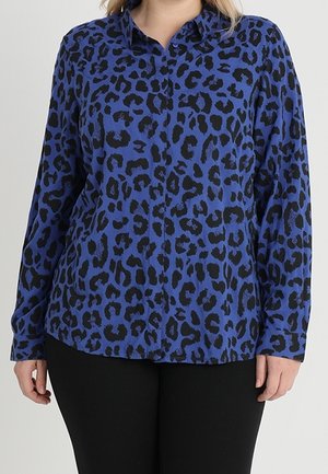 Blouse bleu à manches longues avec imprimé léopard noir, portée avec un pantalon noir, montrant le torse et les bras d'une personne debout de face.
