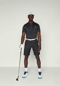 Homem vestindo um conjunto de golfe preto, boné branco, luva e ténis, posicionado com um taco de golfe em frente a um fundo simples.
