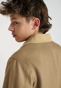 Tan jas met een corduroy kraag, gestructureerde stof en zichtbare stikdetails, gedragen door een persoon die van de achterkant en zijkant te zien is.